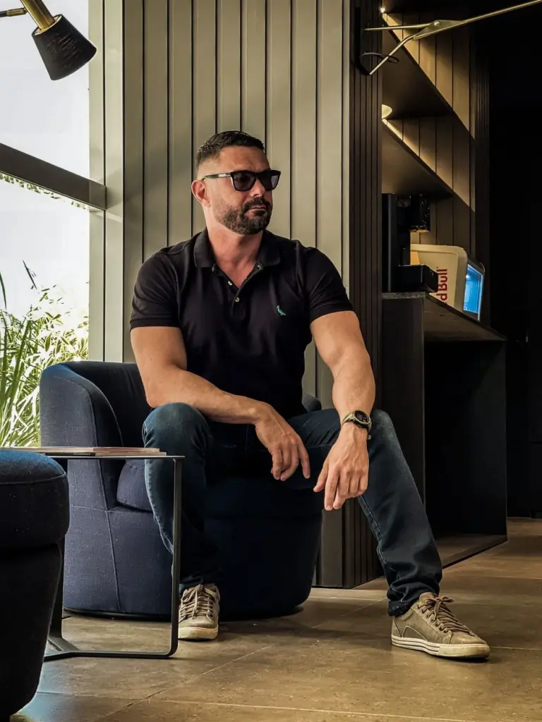 Bearded man in black polo and denim, sitting in a modern room wearing sunglasses.