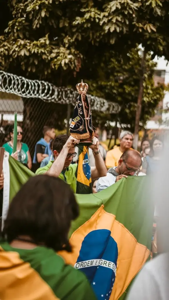 Crowd gathers in Brazil for a religious procession, holding a statue and flags.