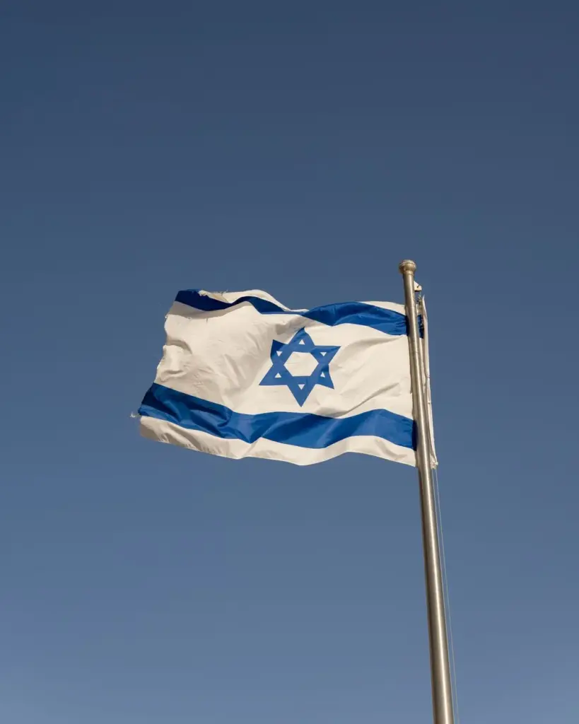 A vibrant Israeli flag on a flagpole waving against a clear blue sky in Jerusalem.