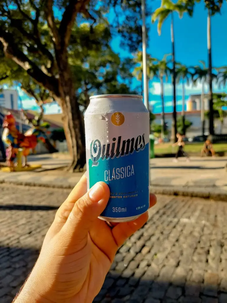Hand holding a cold Quilmes beer can outdoors on a sunny street in Recife, Brazil.