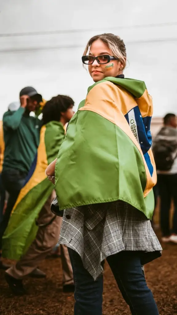 Caucasian woman wearing sunglasses, wrapped in Brazilian flag, attending an outdoor event.