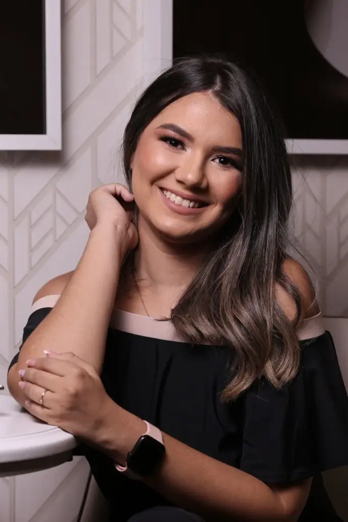 Young female in wearable bracelet looking at camera with toothy smile while touching elbow indoors