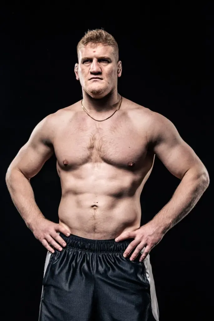 Shirtless bodybuilder showcasing muscular physique with intense expression on dark background.