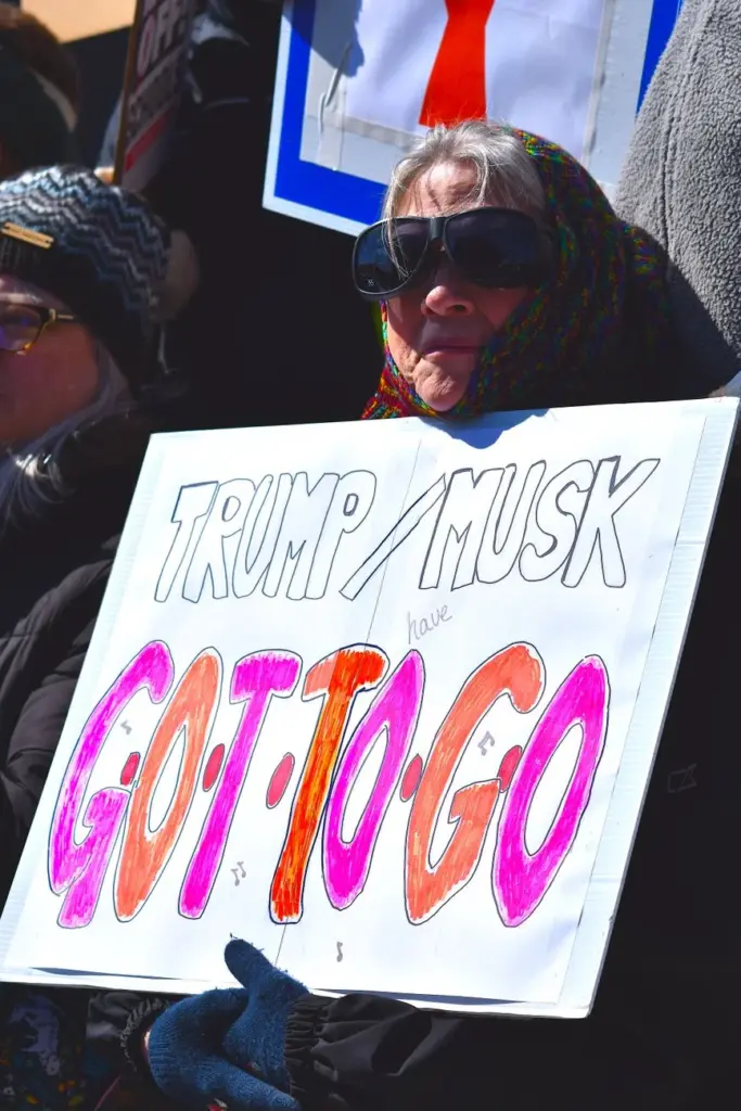 An older woman holds a protest sign against Trump and Musk outdoors in winter.